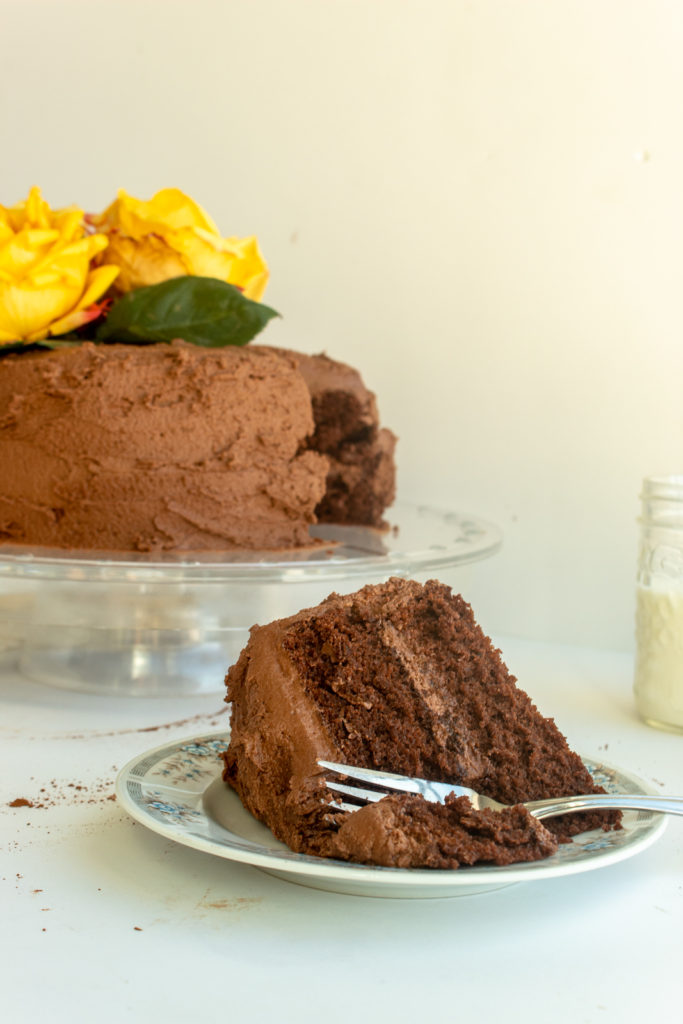 chocolate buttercream frosting on chocolate layer cake on clear cake stand with slice of cake in front straight on shot.