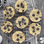 Overhead shot of Chocolate chip protein muffins on round wire rack on wooden surface with extra chocolate chips, glass of milk, wire whisk and teaspoon.