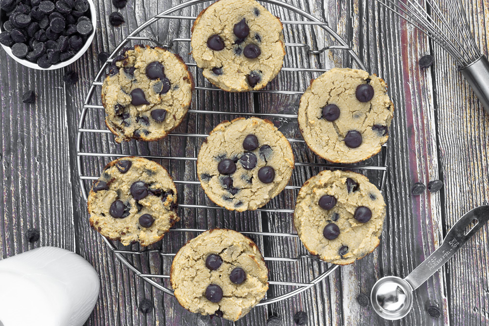 Overhead shot of Chocolate chip protein muffins on round wire rack on wooden surface with extra chocolate chips, glass of milk, wire whisk and teaspoon.