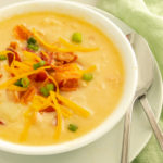 Pressure cooker potato soup in bowl on top of plate with 2 spoons beside green linen.