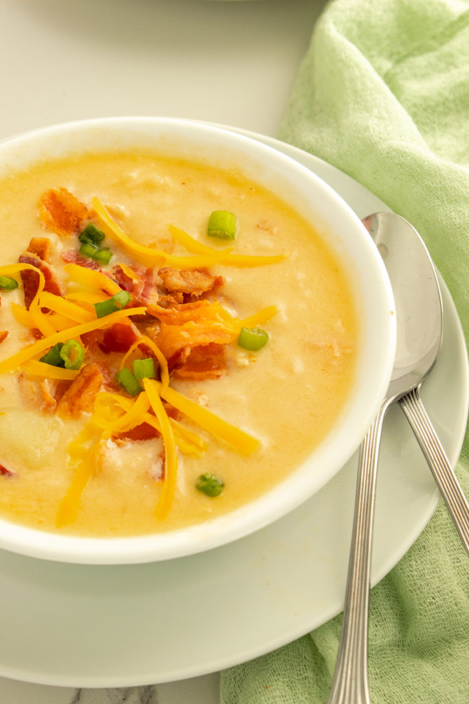 Pressure cooker potato soup in bowl on top of plate with 2 spoons beside green linen.