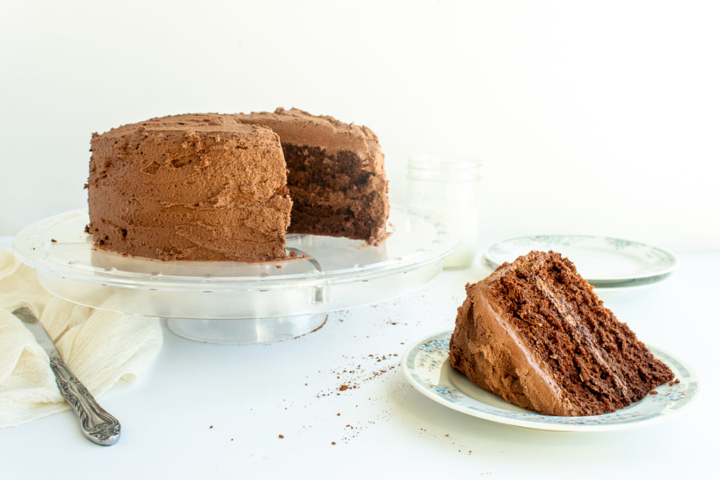 You will be pleased after making this decadent, rich, and moist chocolate layer cake on clear cake stand with piece of cake cut out onto plate.