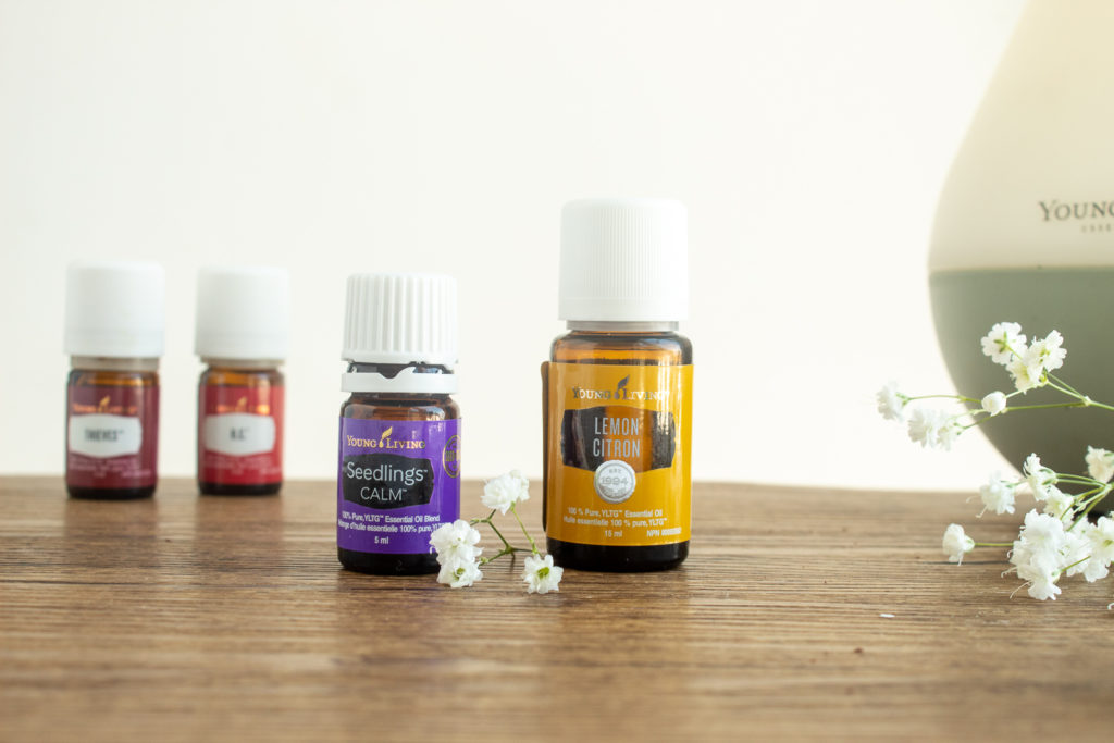 Four Young Living Essential oils on wooden placemat with little white flowers beside dew drop diffuser. Summer 2021 favourites.