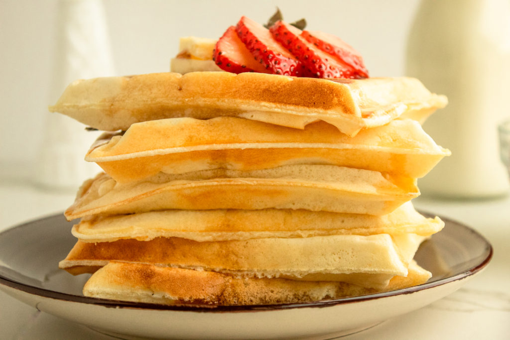 Soft and fluffy waffles stacked on plate with utter and strawberries on top