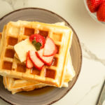 Overhead shot of Soft and fluffy waffles stacked on plate with butter and strawberries on top