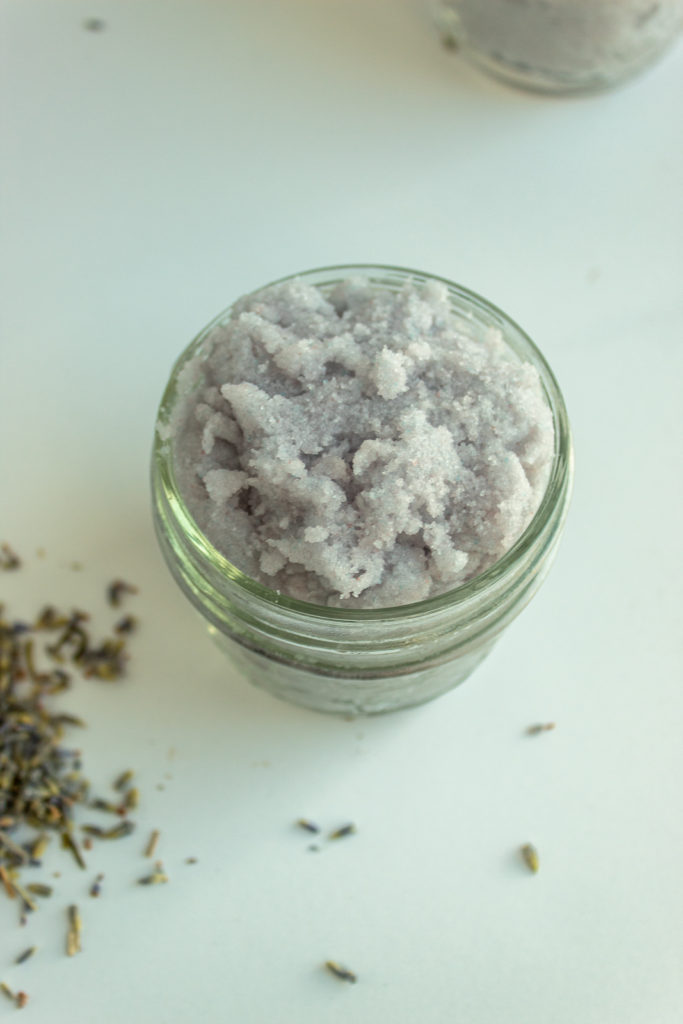 Homemade Lavender Body Scrub in small mason jar next to lavender buds angled shot.