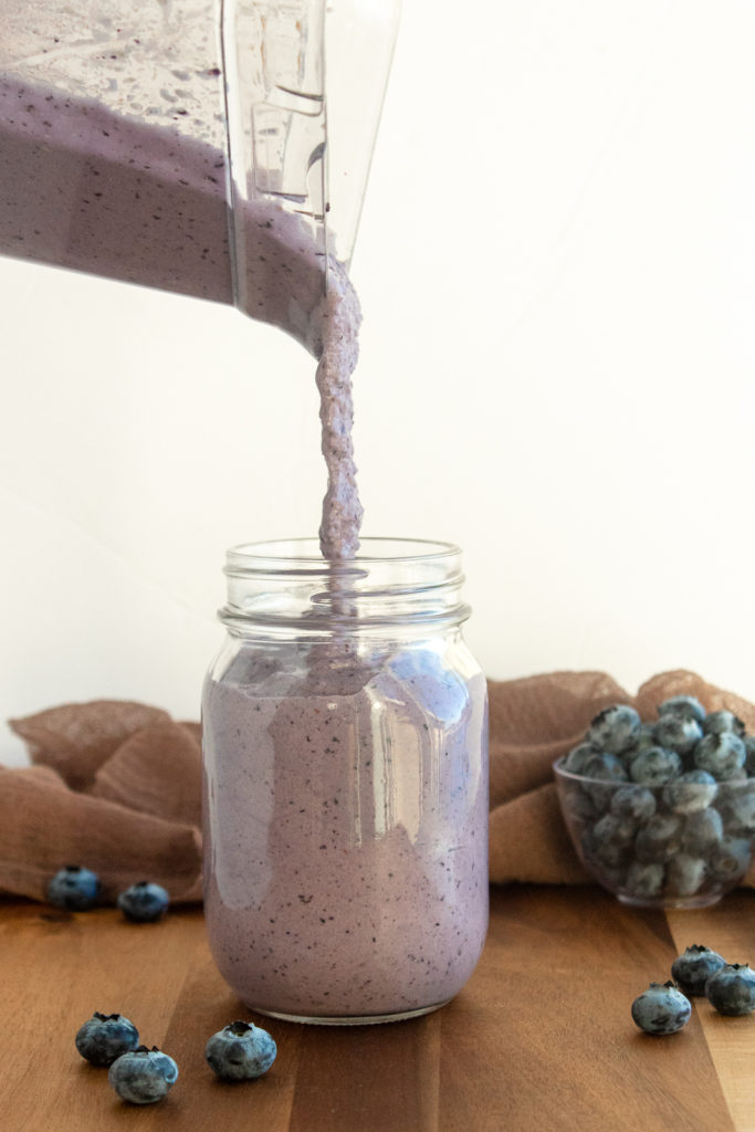 Post workout pb & Jelly Smoothie being poured into glass mason jar on wooden background with blueberries.