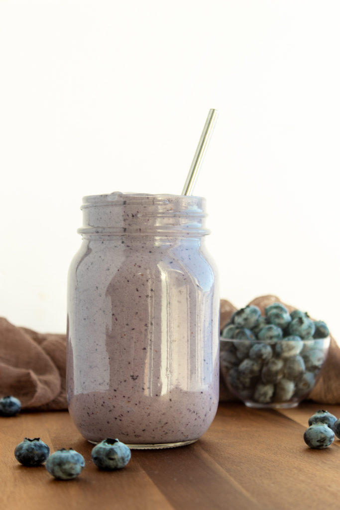 Post workout pb & jelly smoothie in glass mason jar with straw on wooden background with blueberries surrounding.