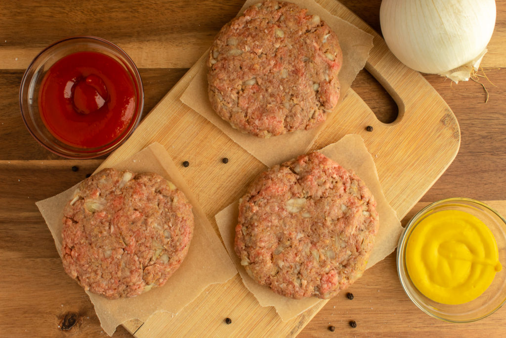 Overhead shot of perfect basic burger patties on cutting board beside bowls of ketchup, mustard, and onion.