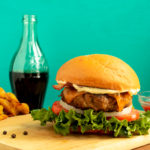 Perfect basic burger on cutting board beside bottle of soda and potato wedges.