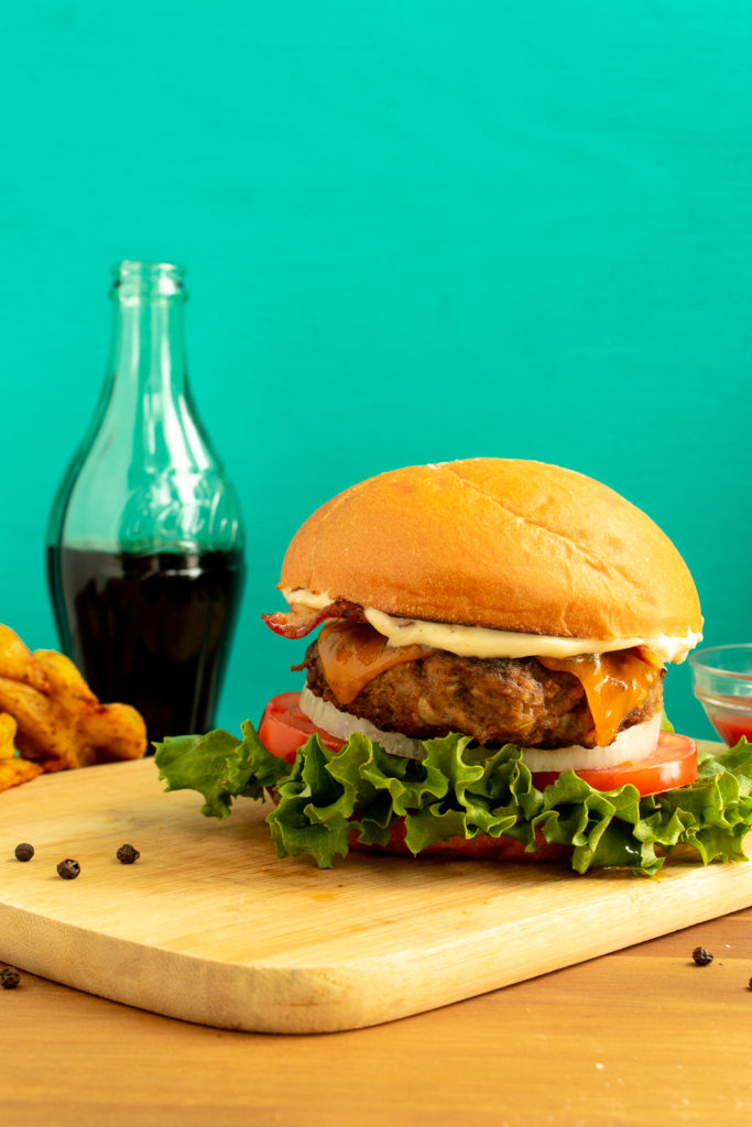 Perfect basic burger on bun with toppings on wooden cutting board beside bowl of ketchup, coke, and wedges. 