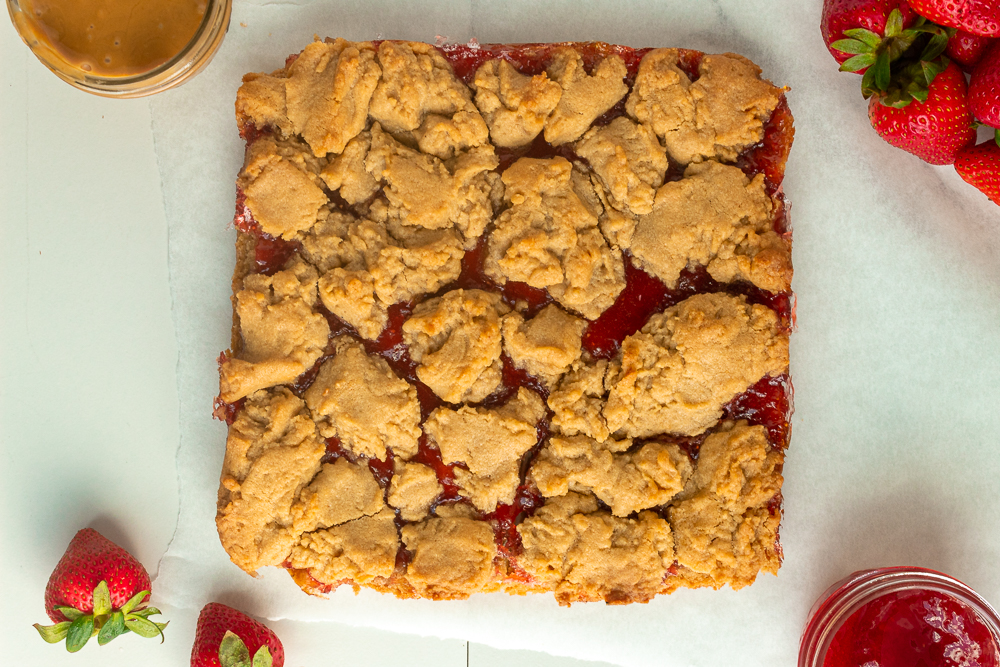 Peanut butter & jelly bars uncut on parchment paper overhead shot with strawberries, and jars of peanut butter and jam surrounding. 