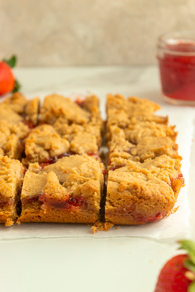 Peanut butter & jelly bars on parchment paper angled shot with strawberries and jam surrounding.