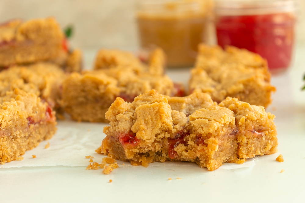 Peanut butter & jelly bars on parchment paper straight shot with peanut butter and jam in background.