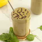 Chocolate Protein Green Smoothie on wooden coaster surrounded by spinach and a banana and extra smoothie in background angled shot.