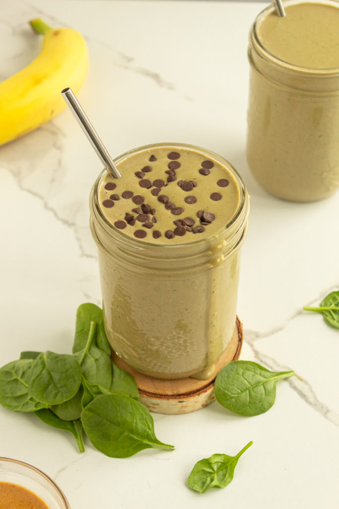 Chocolate Protein Green Smoothie on wooden coaster surrounded by spinach and a banana and extra smoothie in background angled shot.