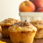 Peach Streusel Muffins on wooden cutting board with tray of peaches in background.