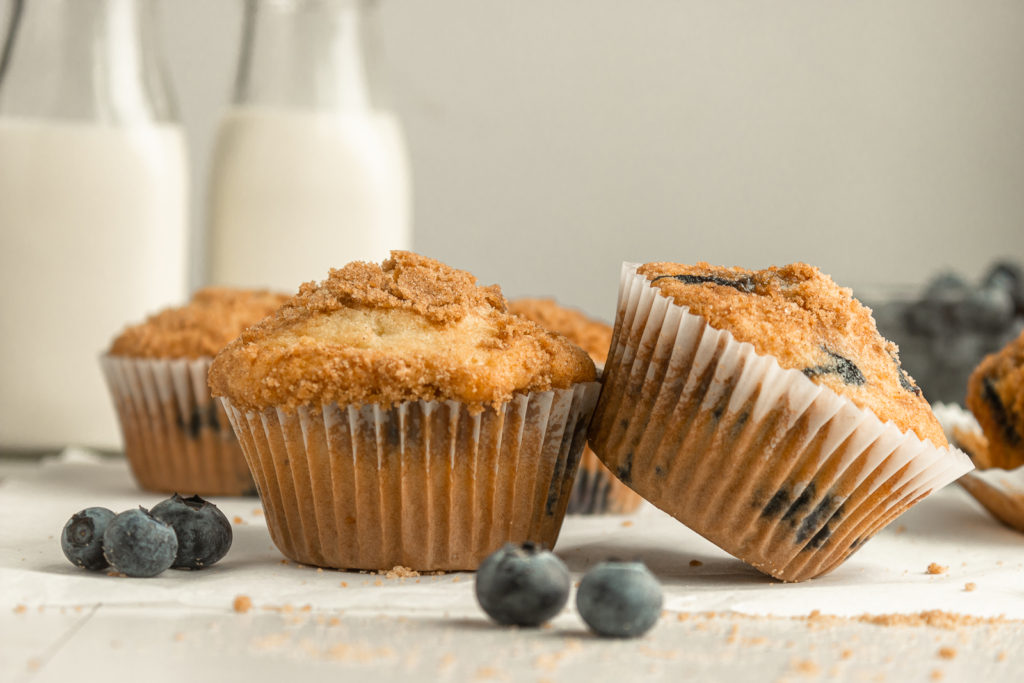 Straight on shot of blueberry streusel muffins with paper cups with white backgrounds and surrounded by blueberries.