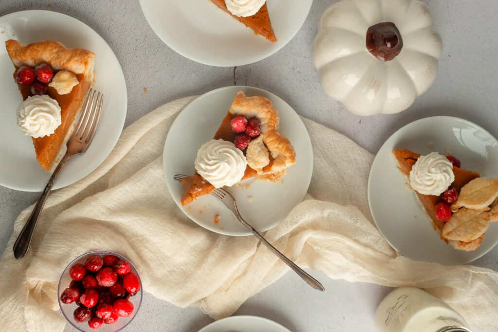 Overhead shot of individual slices of pumpkin pie on white plates with whipped cream, sugared cranberries and forks.