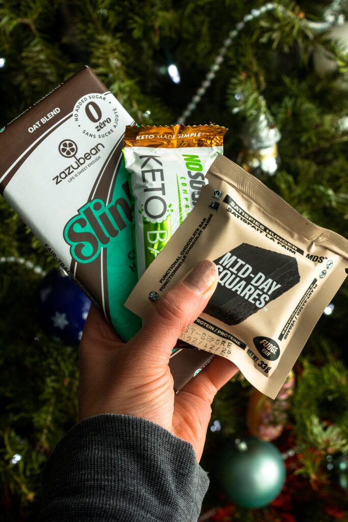 Slinky, keto bar, and fudge midday square held in hand in front of Christmas tree.