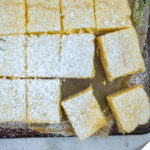 Overhead shot of Easy Lemon bars cut into squares on parchment on pan with purple linen on side and yellow flower on top.