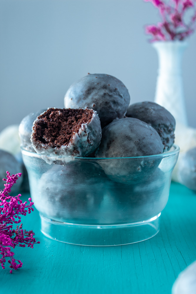 Straight on shot of chocolate glazed donut holes in clear bowl. A bite taken out of one. Purple floral in background and foreground.