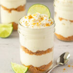 Chilled no-bake key lime pie mousse cups garnished with lime slices and crushed graham crackers with spoon beside.