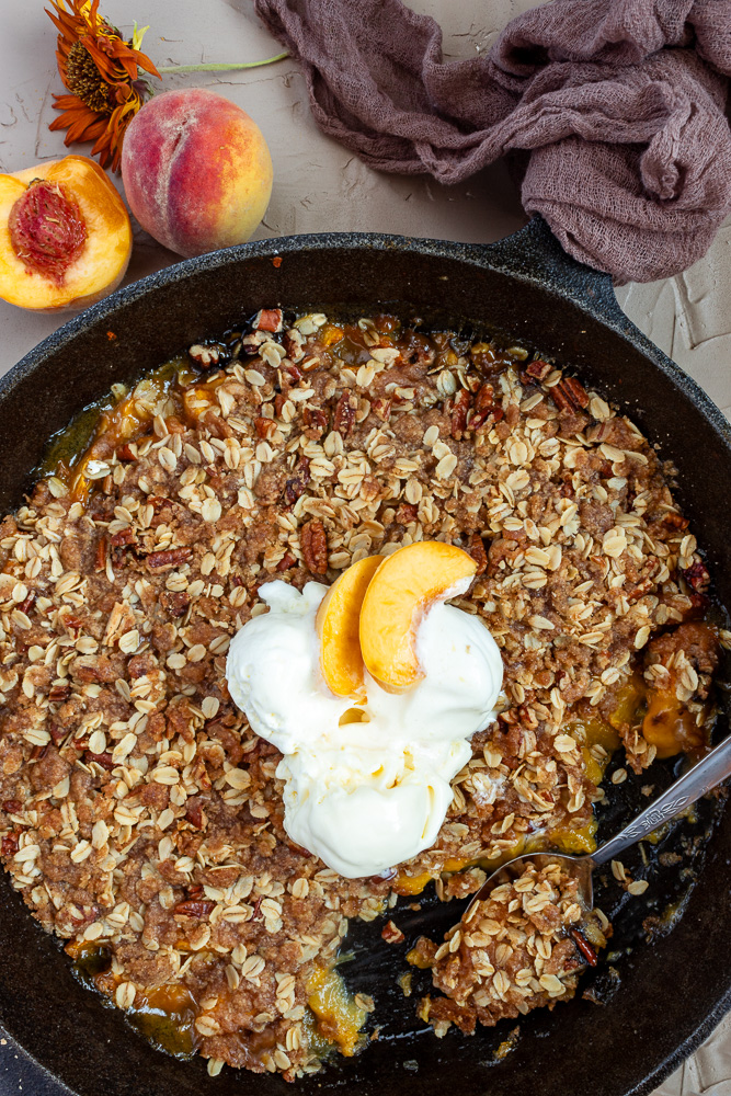 Overhead shot of peach crisp in cast iron skillet with vanilla ice cream and spoon. 