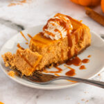 Slice of pumpkin cheesecake with gingerbread crust and topped with whipped cream and chai caramel sauce.