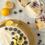 Overhead shot of simple lemon blueberry cake on cake stand with 3 slices on plates beside. Surrounded by some lemons and blueberries.