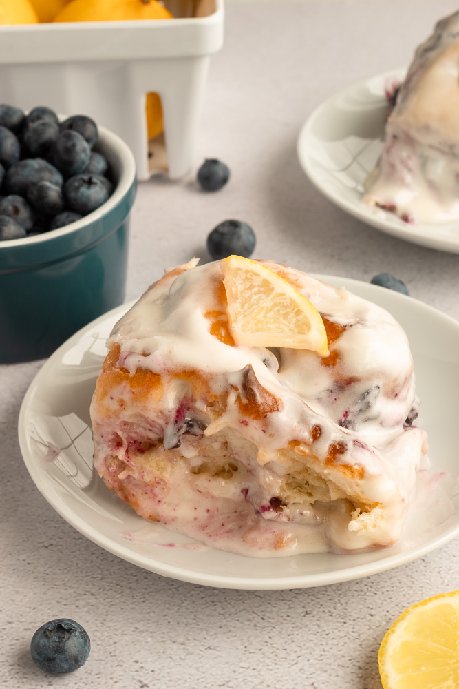Lemon Blueberry Sweet Roll on plate agnled shot surrounded by lemons and blueberries.